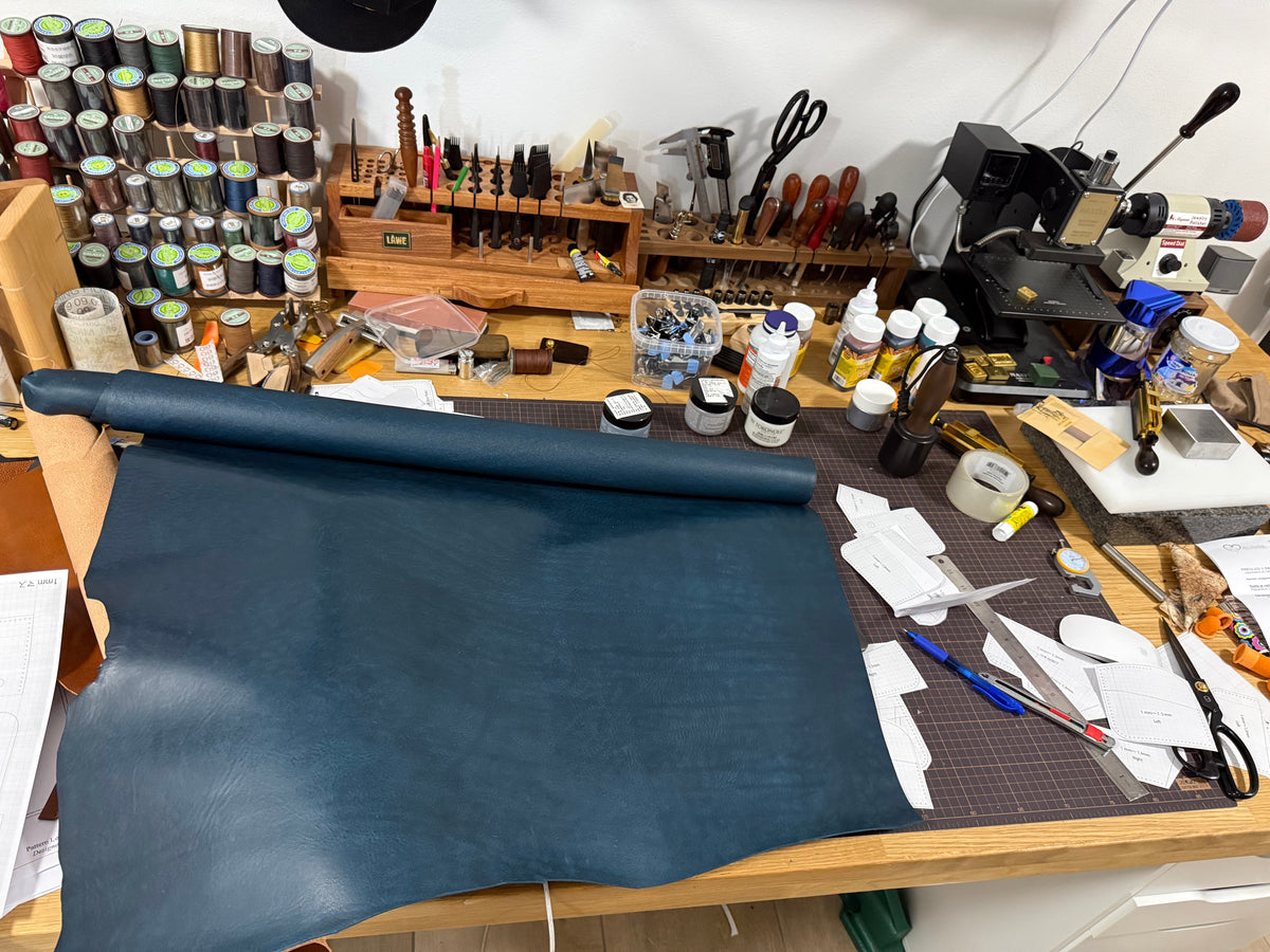 LÅWE workshop bench with a navy full-grain leather hide unrolled across it, backed by racks of thread and hand tools