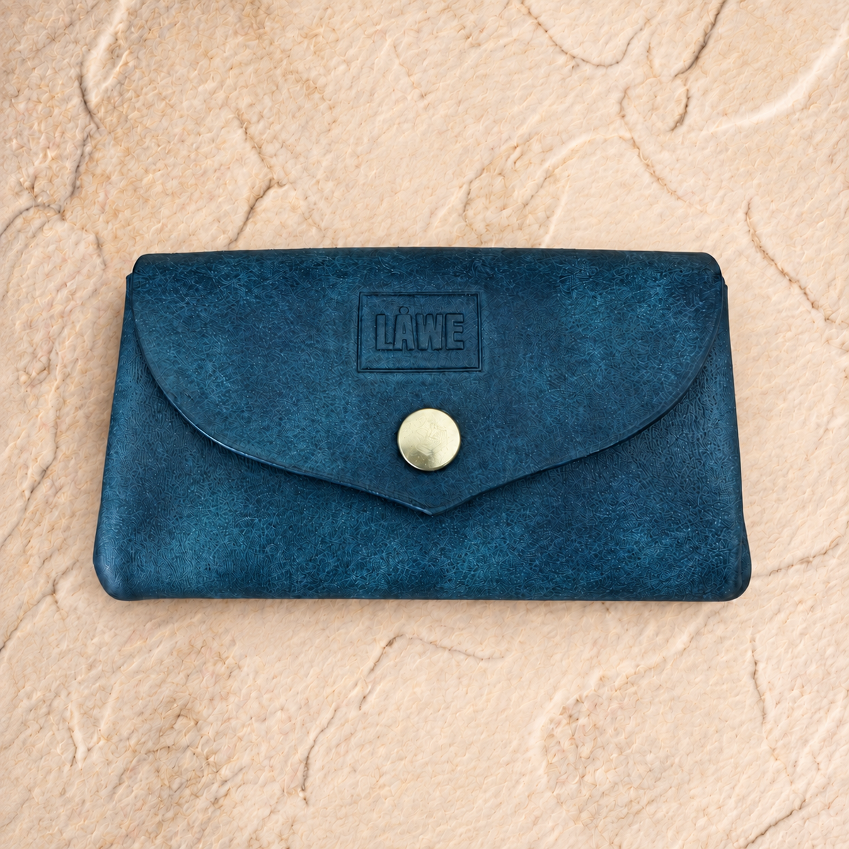 A dark blue textured full-grain leather wallet with a flap closure and a gold snap button, featuring an embossed 'LÅWE' logo on the front, set against a beige stone background.