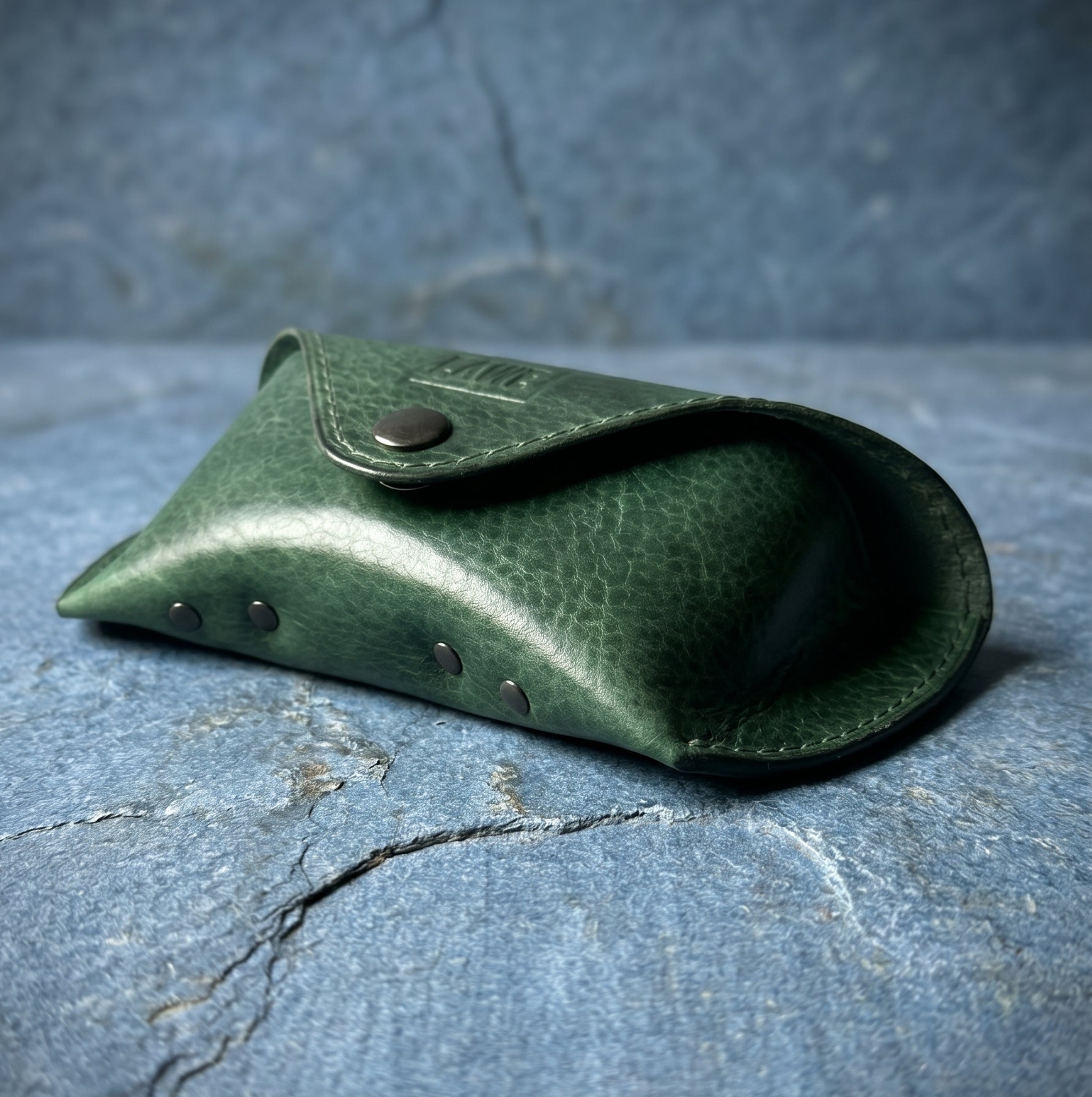 Green leather eyeglasses case with metal studs on a textured blue stone surface