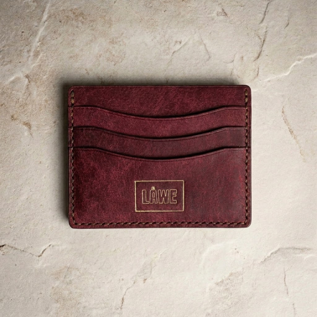 A bordeaux leather card holder wallet with a debossed square logo patch reading 'LÅWE' on the front, displayed flat on a textured beige stone surface.