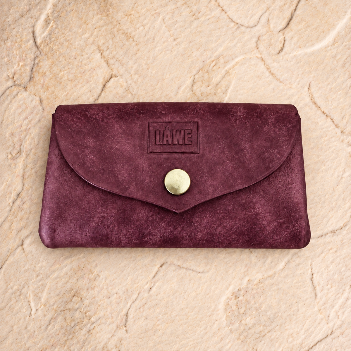 A burgundy natural leather envelope-style wallet with a snap button closure and an embossed 'LÅWE' logo, displayed on a textured beige stone surface.