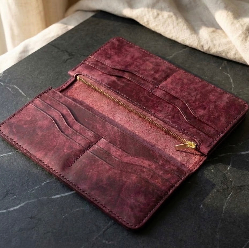 A dark burgundy distressed leather wallet shown open, featuring multiple card slots, a zippered compartment, and a gold zipper pull, resting on a dark gray marble surface with a beige fabric background.