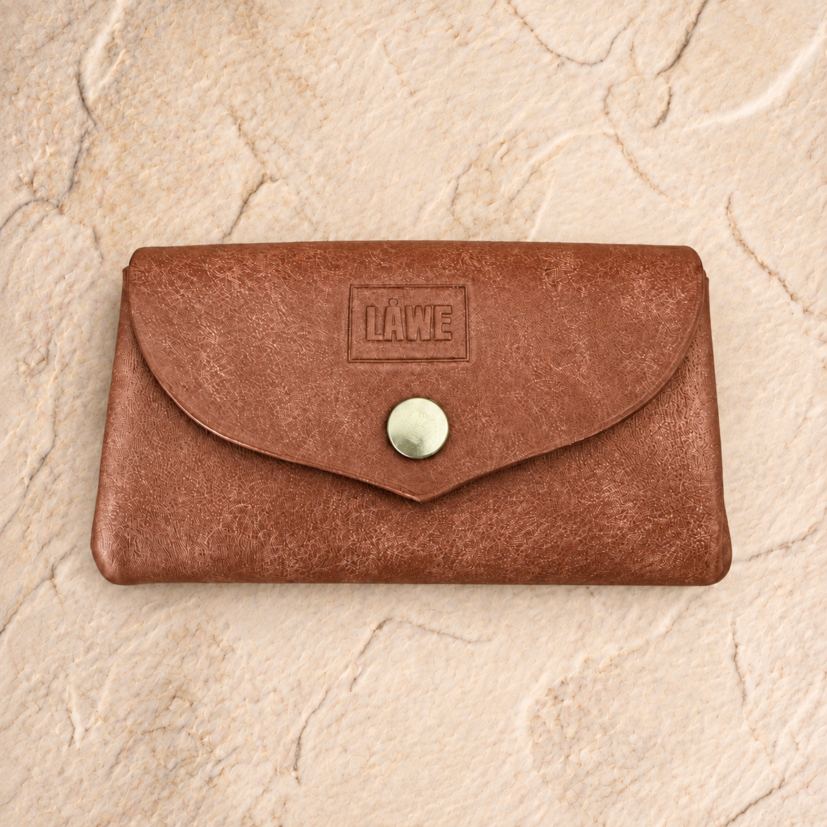 A brown leather wallet with a flap closure and a golden snap button, featuring an embossed 'LÅWE' logo on the front, set against a textured beige stone background.