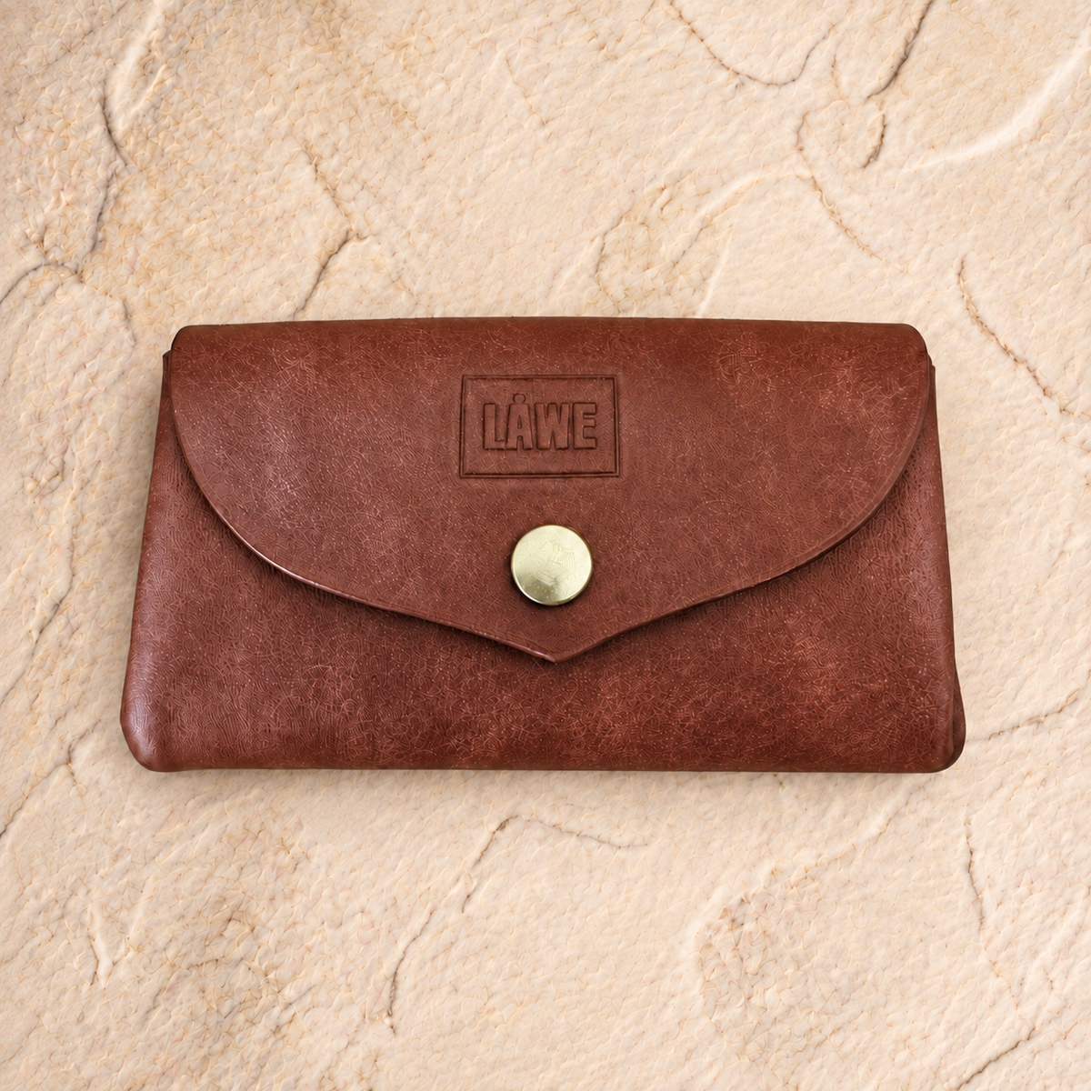 A brown leather wallet with a flap closure and a round snap button, featuring an embossed 'LÅWE' logo on the front, displayed on a textured beige stone surface.