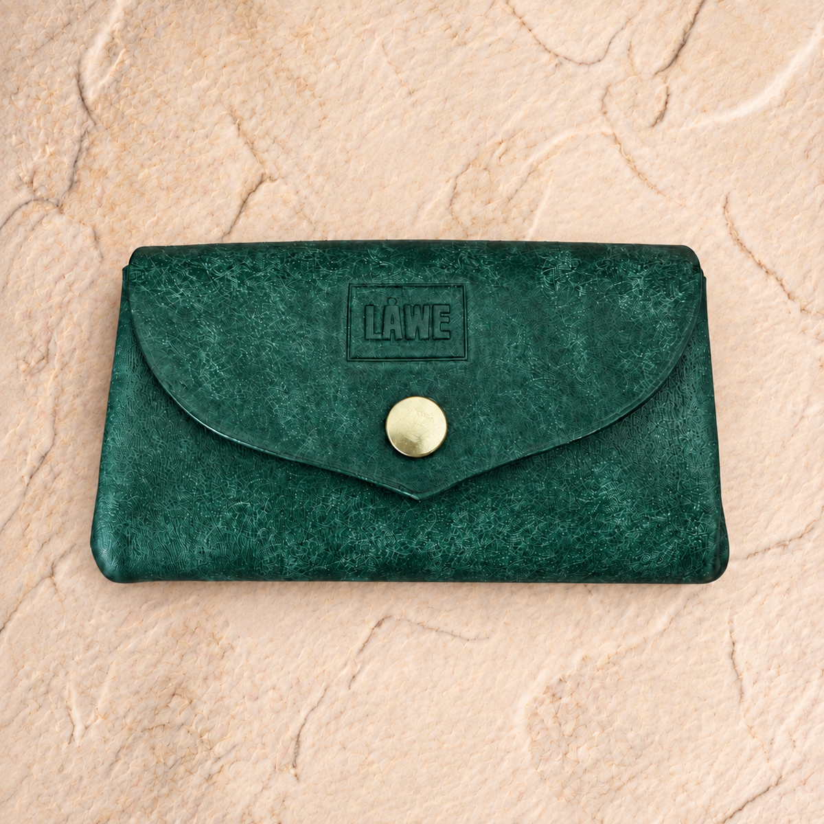 A dark green textured leather wallet with a flap closure and a gold snap button, featuring an embossed 'LÅWE' logo on the front, set against a beige stone background.