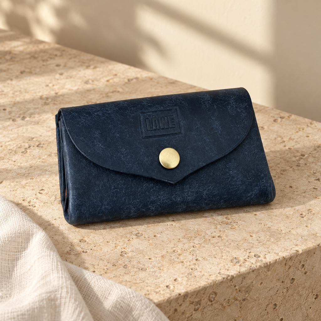 A dark navy blue textured wallet with a gold snap button and a debossed 'LÅWE' logo on the front flap, resting on a beige stone surface with soft natural shadows.