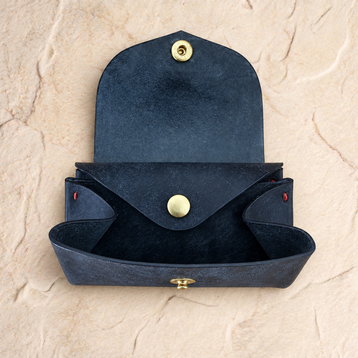 A dark navy blue textured leather pouch with gold-tone metal snap buttons, shown open against a textured beige stone background.