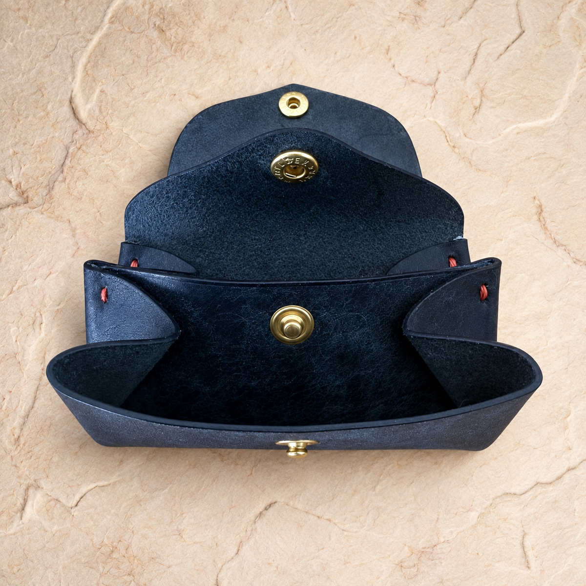 A dark navy blue leather clutch wallet with gold-tone snap buttons, shown open against a textured beige stone background.