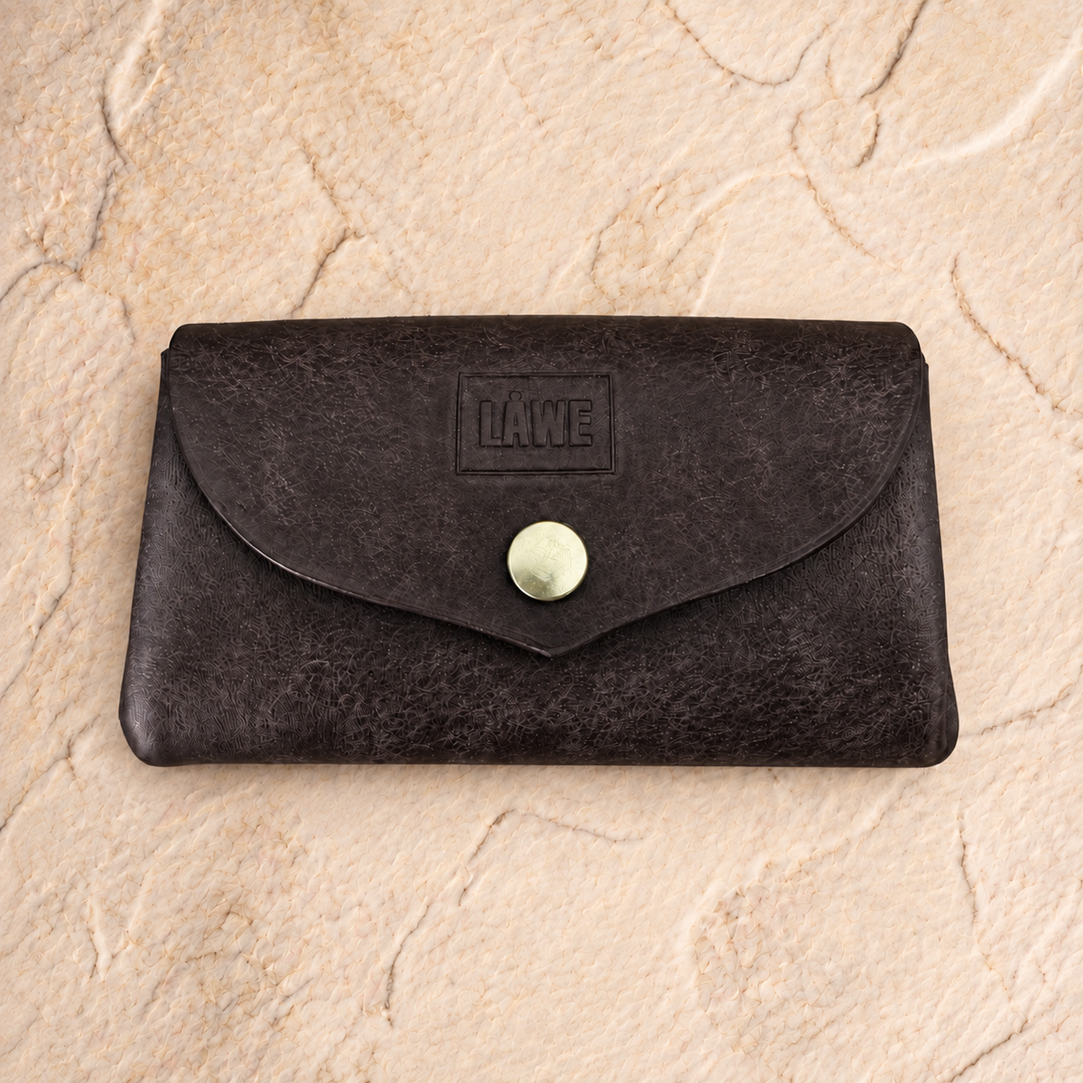 A dark brown textured leather wallet with a flap closure and a round metal snap button, featuring an embossed 'LÅWE' logo on the front, set against a light beige stone background.