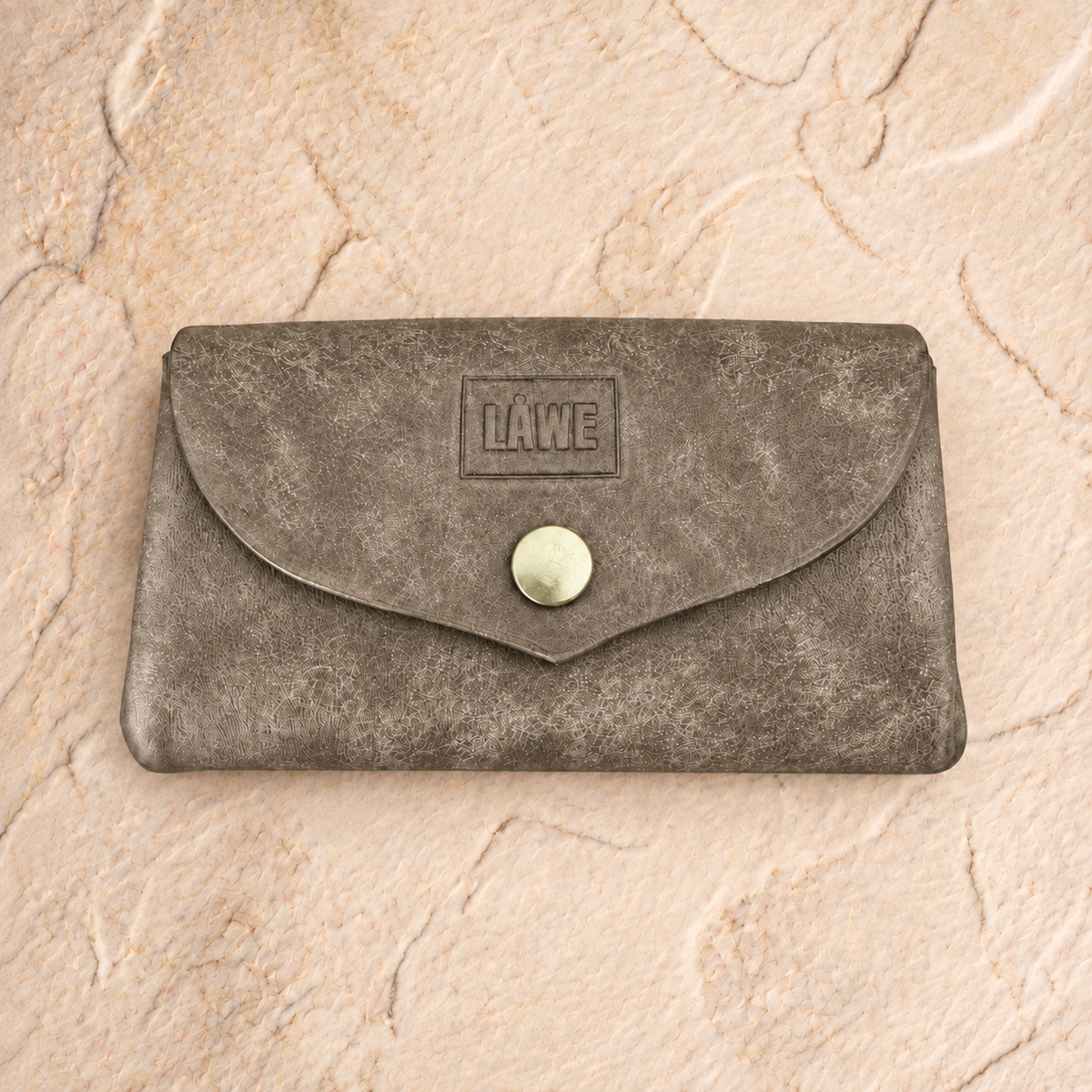 A small gray textured leather pouch with a flap closure and a round metal snap button, featuring an embossed 'LÅWE' logo on the front, displayed on a beige stone surface.