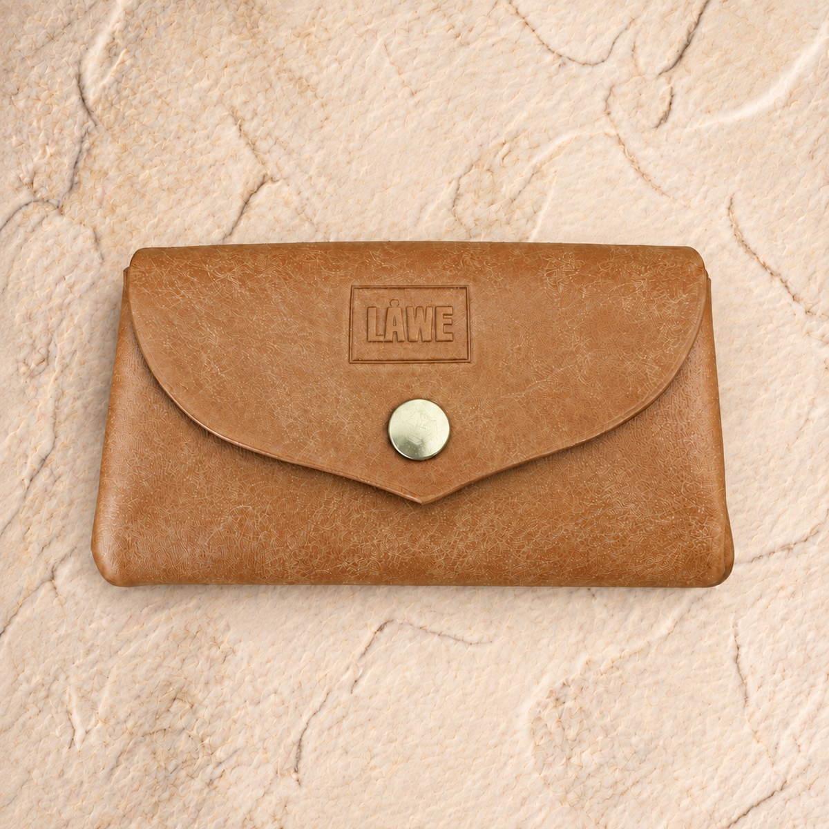 A light brown leather card holder wallet with a flap closure and a golden snap button, featuring an embossed 'LÅWE' logo on the front, displayed on a textured beige stone surface.