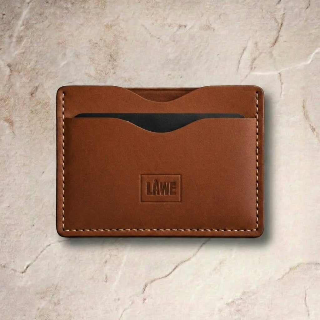 A brown leather card holder wallet with visible stitching and a debossed 'LÅWE' logo on the front, displayed on a textured beige stone surface.