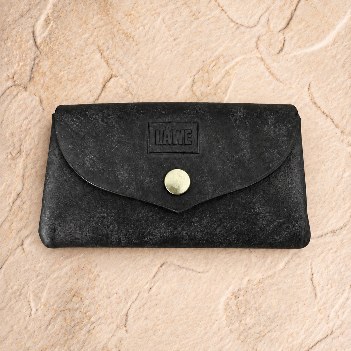 A black textured leather wallet with a flap closure and a gold snap button, featuring an embossed 'LÅWE' logo, displayed on a beige stone background.