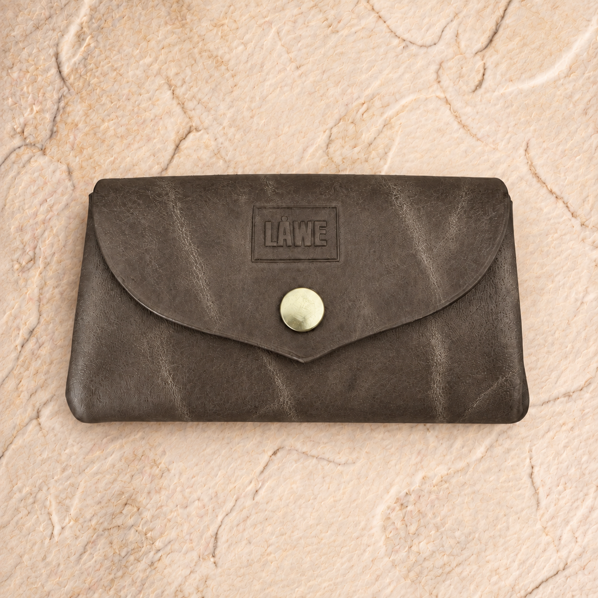 An olive grey leather wallet with a flap closure and a round snap button, featuring an embossed 'LÅWE' logo on the front, displayed on a textured beige stone surface.