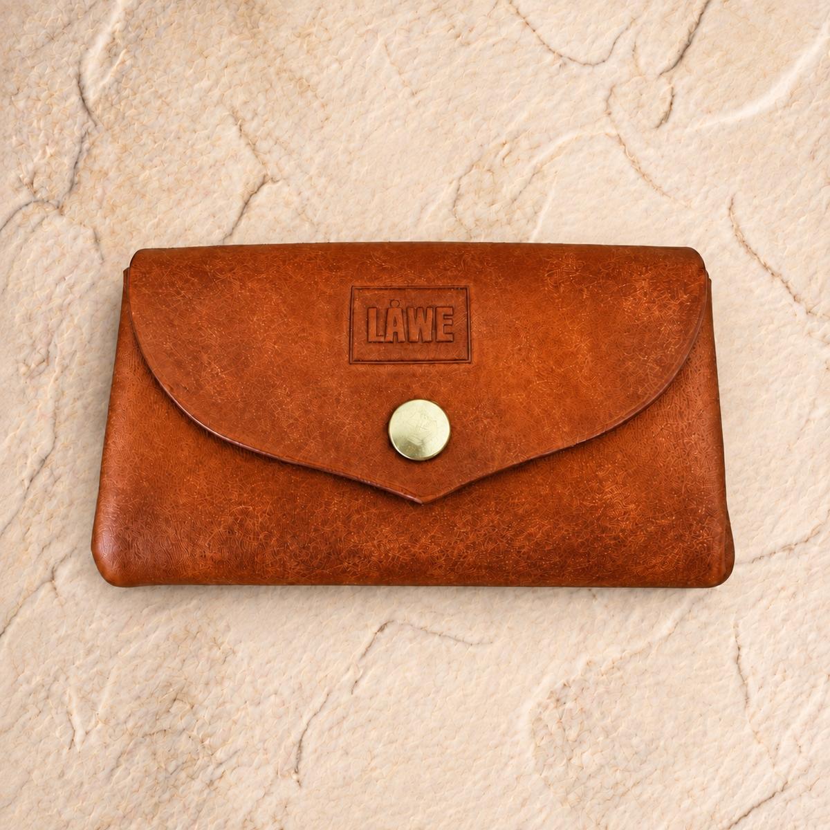 A brown leather wallet with a flap closure and a brass snap button, featuring an embossed 'LÅWE' logo on the front, set against a textured beige stone background.