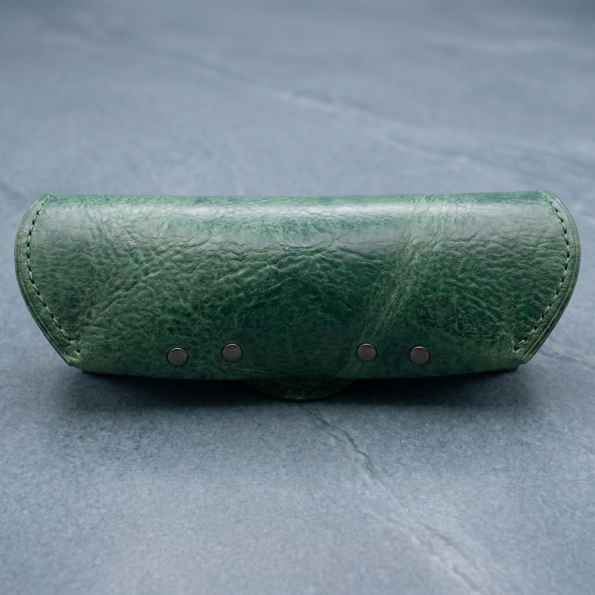 Green leather eyeglasses case with four small metal rivets on a grey background