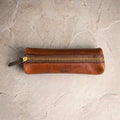 LÅWE Trace leather pen case in Whiskey, shown closed with a gold zipper and a small embossed 'LÅWE' logo, placed on a light beige stone surface.
