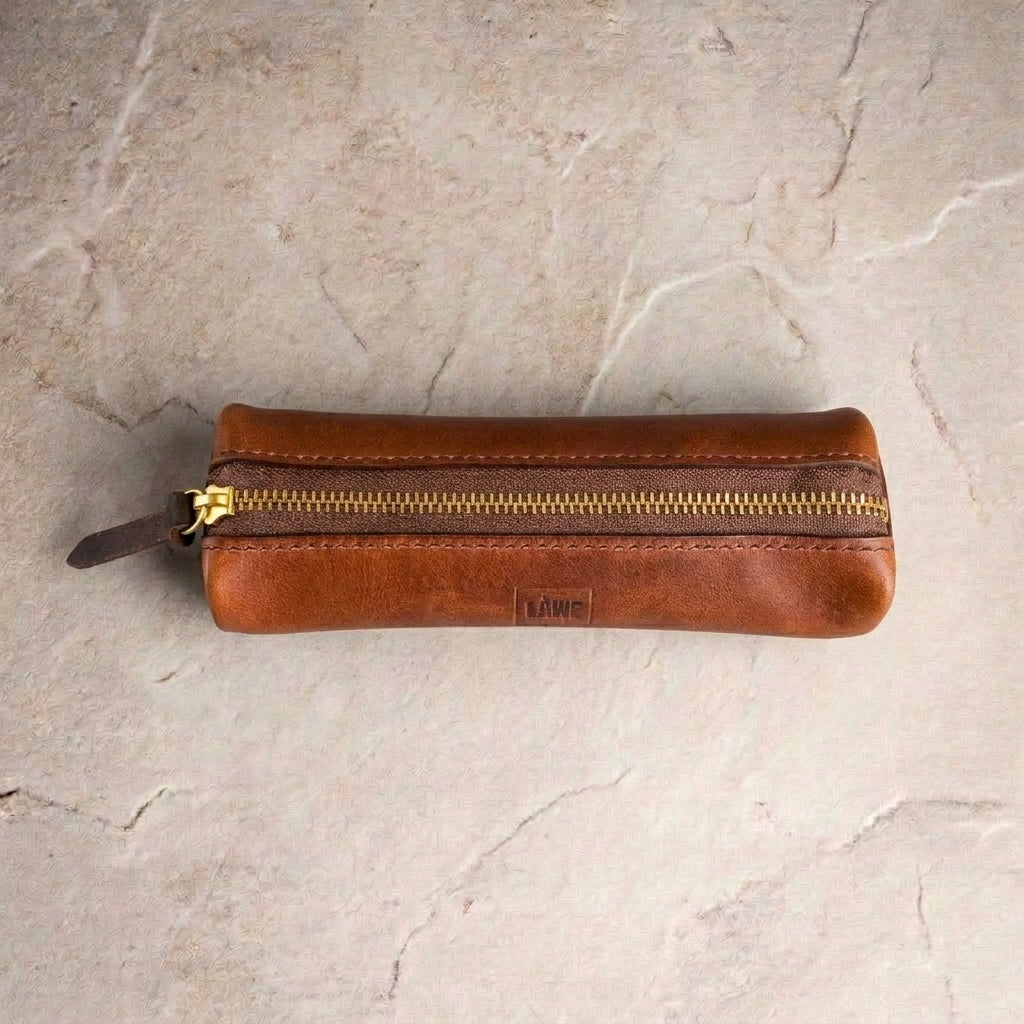 LÅWE Trace leather pen case in Whiskey, shown closed with a gold zipper and a small embossed 'LÅWE' logo, placed on a light beige stone surface.