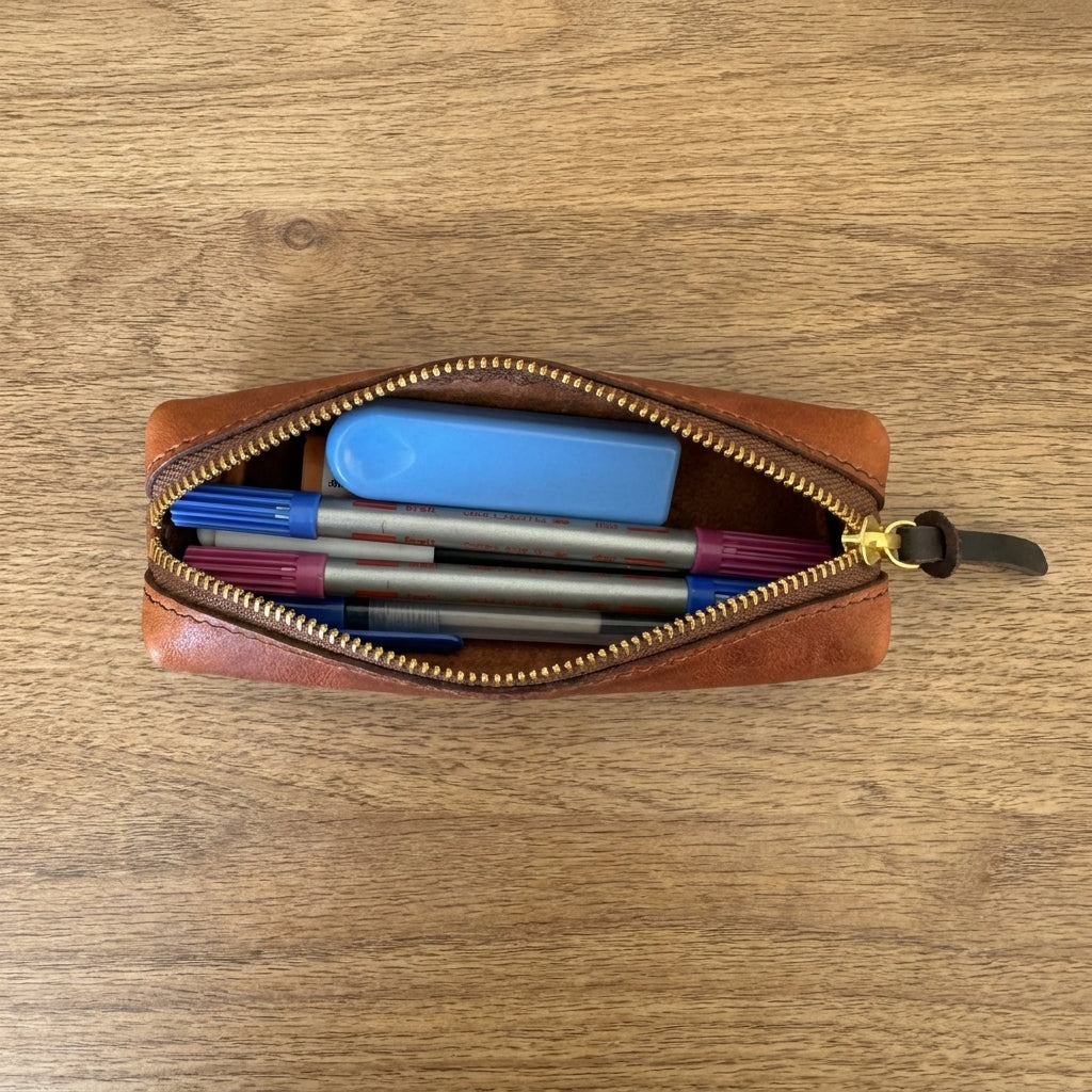 A brown leather zippered pencil case, shown open from a top-down view on a wooden table. Inside, it is filled with various pens and a blue eraser.
