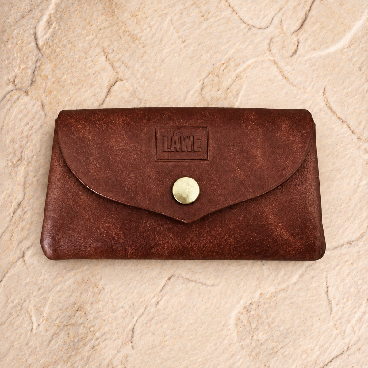 A brown leather wallet with a flap closure and a gold snap button, featuring an embossed 'LÅWE' logo on the front, displayed on a textured beige stone surface.
